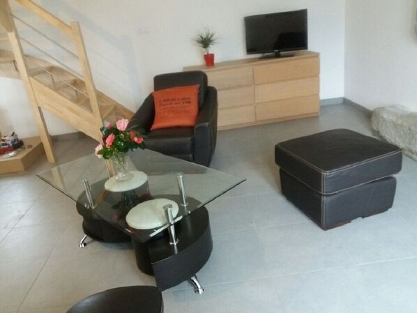 A modern living room with a glass coffee table, a black sofa with an orange cushion, a black ottoman, a wooden TV stand, and a staircase. - A vacation rental between the sea and the countryside   (Sibiril)