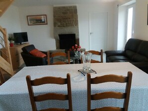 A dining area with a table set for two, surrounded by wooden chairs. A fireplace is visible in the background, and a television is placed on a wooden cabinet to the left. - A vacation rental between the sea and the countryside   (Sibiril)
