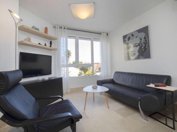 Ein modernes Wohnzimmer mit Flachbildfernseher, einem schwarzen Ledersofa, einem schwarzen Leder-Sessel, einem kleinen runden Couchtisch und einem großen Fenster mit Vorhängen.