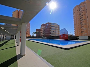 A swimming pool with a clear blue water surface, surrounded by green artificial turf and white tiled flooring. In the background, there are multi-story residential buildings with balconies.