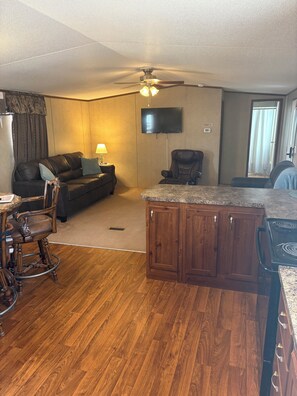A living room with a sofa, a television, a ceiling fan, and a kitchen area with a stove.