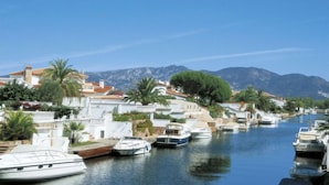 Un port de plaisance où des bateaux sont amarrés le long d’un canal, entouré de bâtiments et avec des montagnes en arrière-plan.