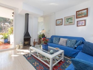 A living room with a blue sofa, a glass coffee table, and a fireplace. There are three framed pictures on the wall and a window with a view of greenery.