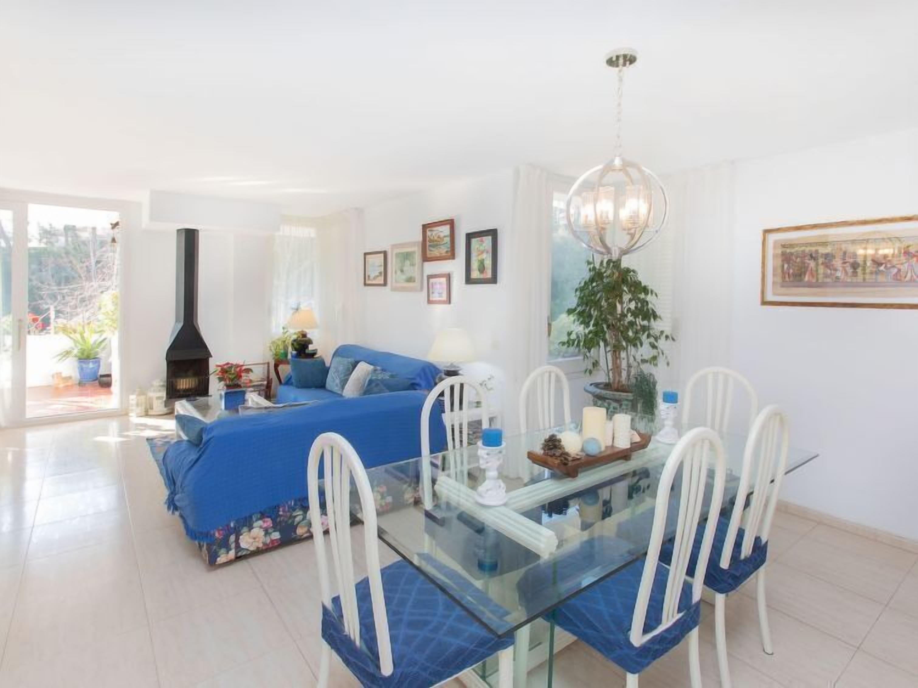 Un salon lumineux avec un canapé bleu, une table à manger en verre et des chaises blanches, une cheminée noire et un lustre.
