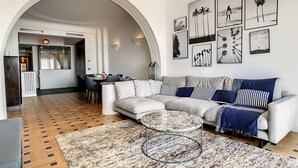 A modern living room with a sectional sofa, a coffee table, and a patterned rug. There are framed pictures on the wall and a dining area in the background. - N&J - BLUE PROMENADE - Sea front - central - Luxe (Niza)