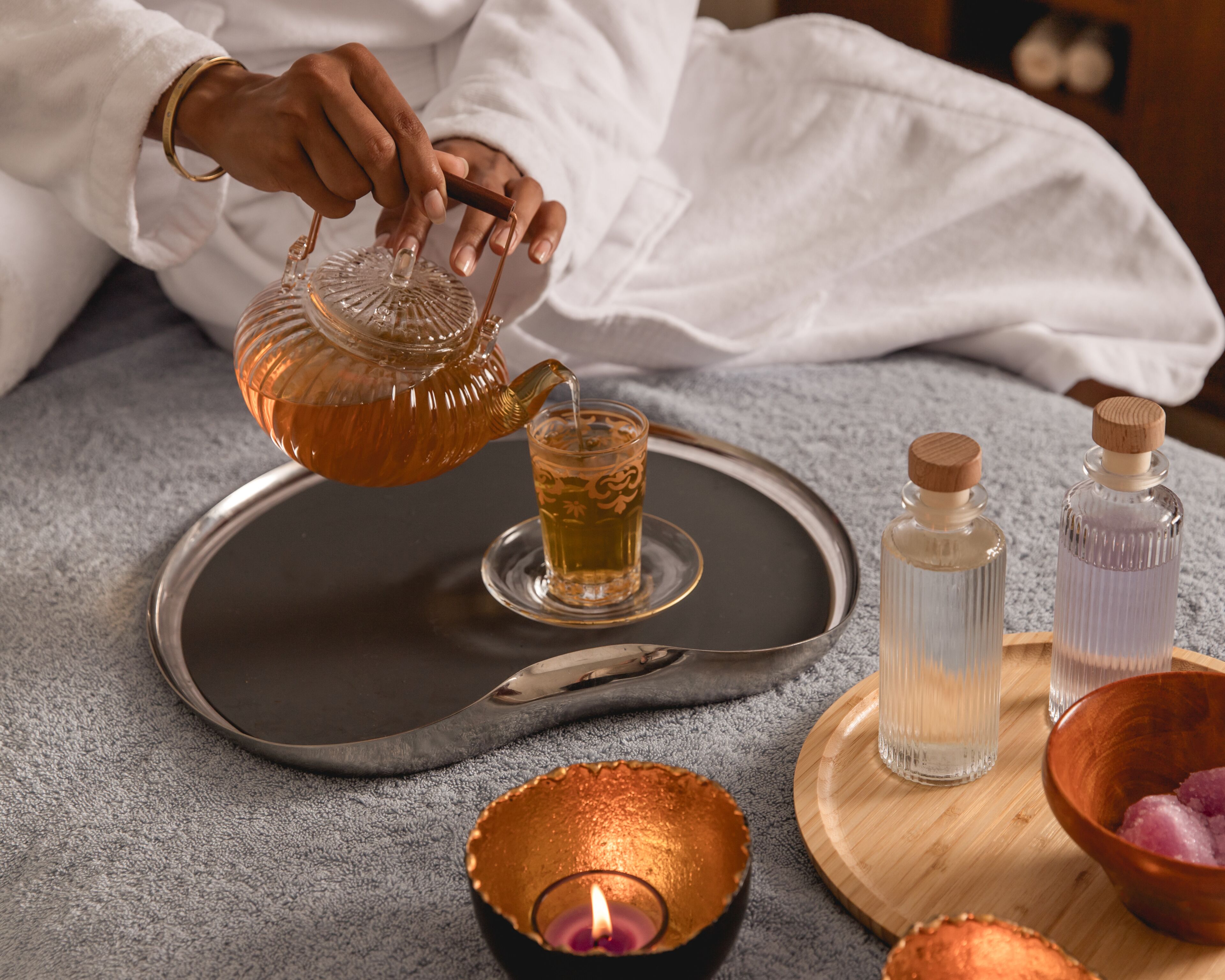 A person pouring tea into a glass, with a tray, candles, and small bottles on a table.