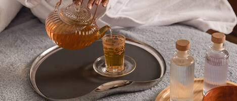A person pouring tea into a glass, with a tray, candles, and small bottles on a table.