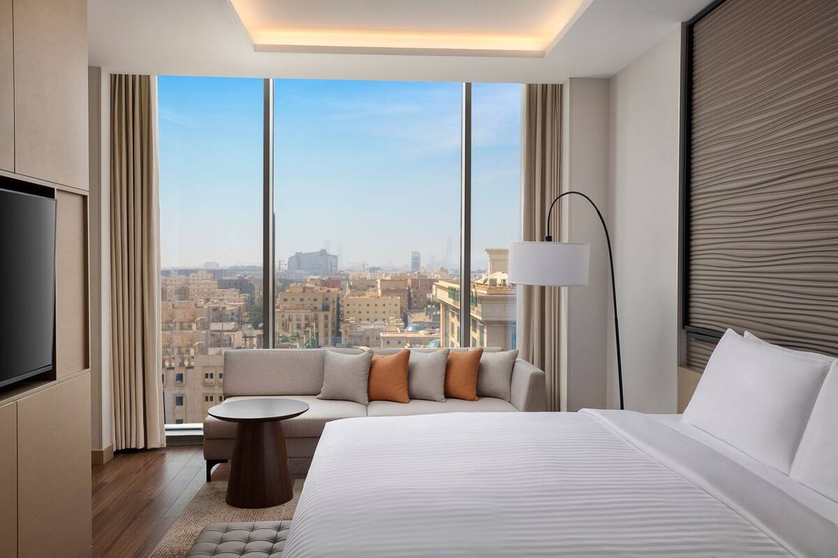 Une chambre d’hôtel moderne, dotée d’un grand lit, d’un canapé, d’une petite table ronde et d’un téléviseur. La chambre offre une vue sur la ville grâce à de grandes fenêtres.