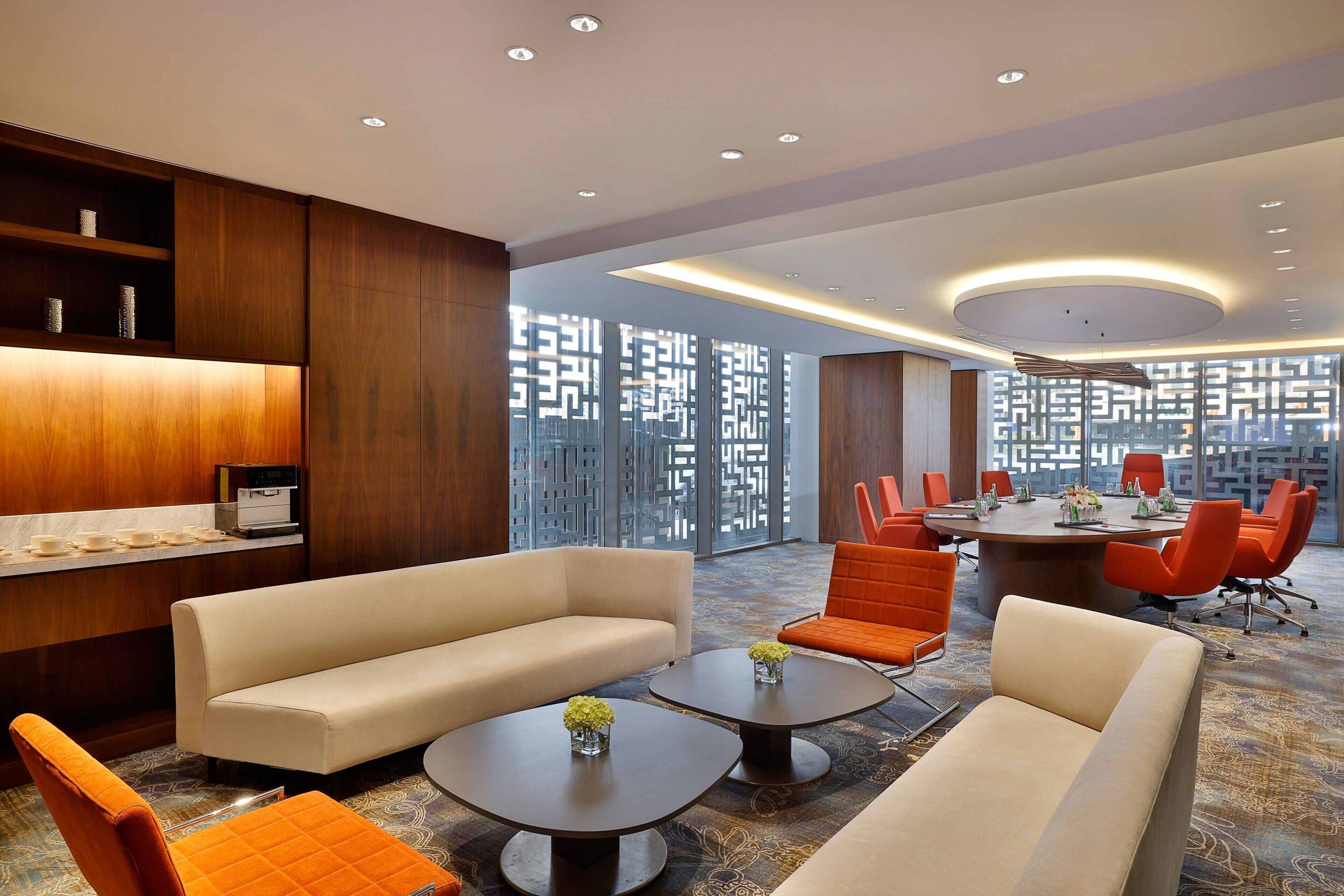 Un moderno vestíbulo de hotel con un sofá, mesas auxiliares y sillas color naranja. Hay una mesa de conferencias con sillas y una pared decorativa con patrones geométricos.
