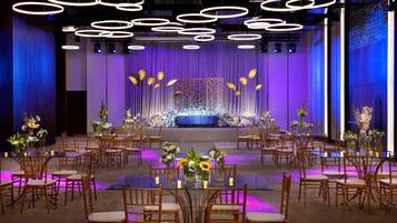 A banquet hall with round tables, chairs, and a stage with floral arrangements.