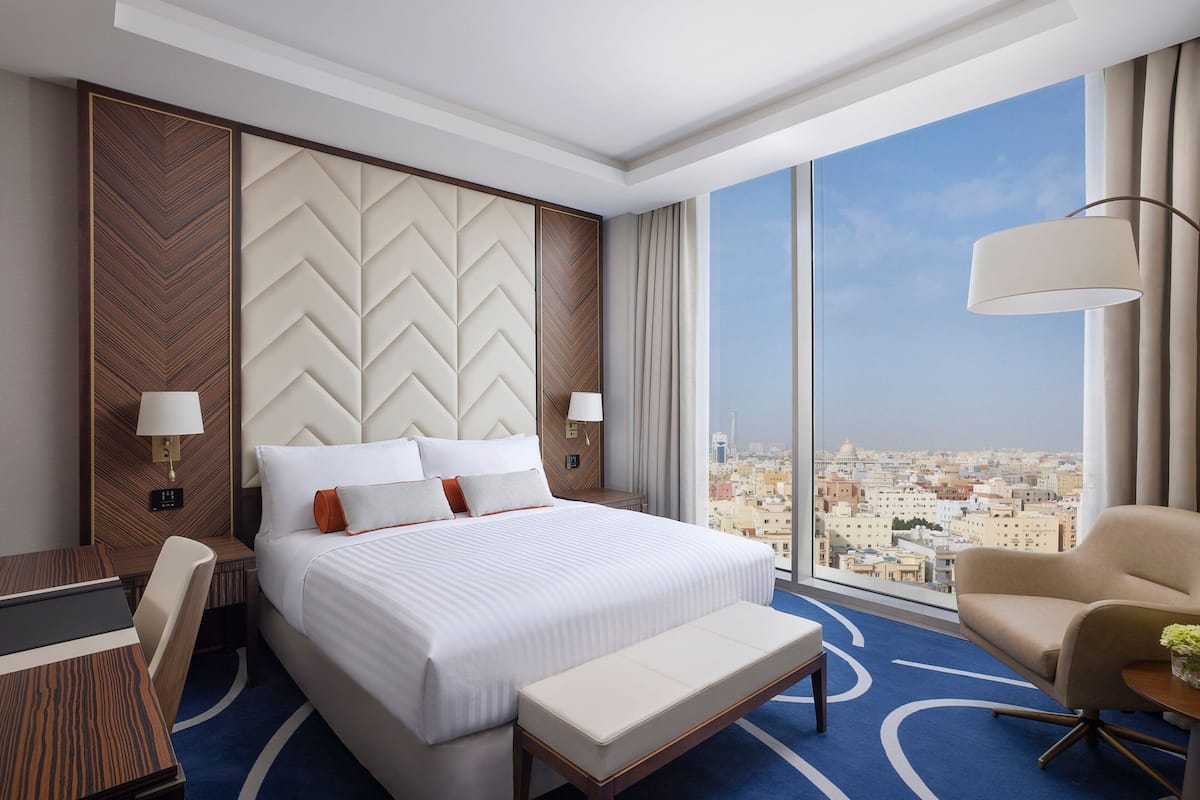 Une chambre d’hôtel dotée d’un grand lit, d’un bureau, d’une chaise, d’un banc et offrant une vue sur la ville grâce à de grandes fenêtres.