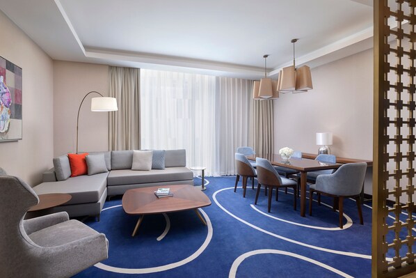Una habitación de hotel moderna con sofá modular, una mesa de centro redonda, zona de comedor con sillas y mesa, un ventanal con cortinas y un panel decorativo en la pared.
