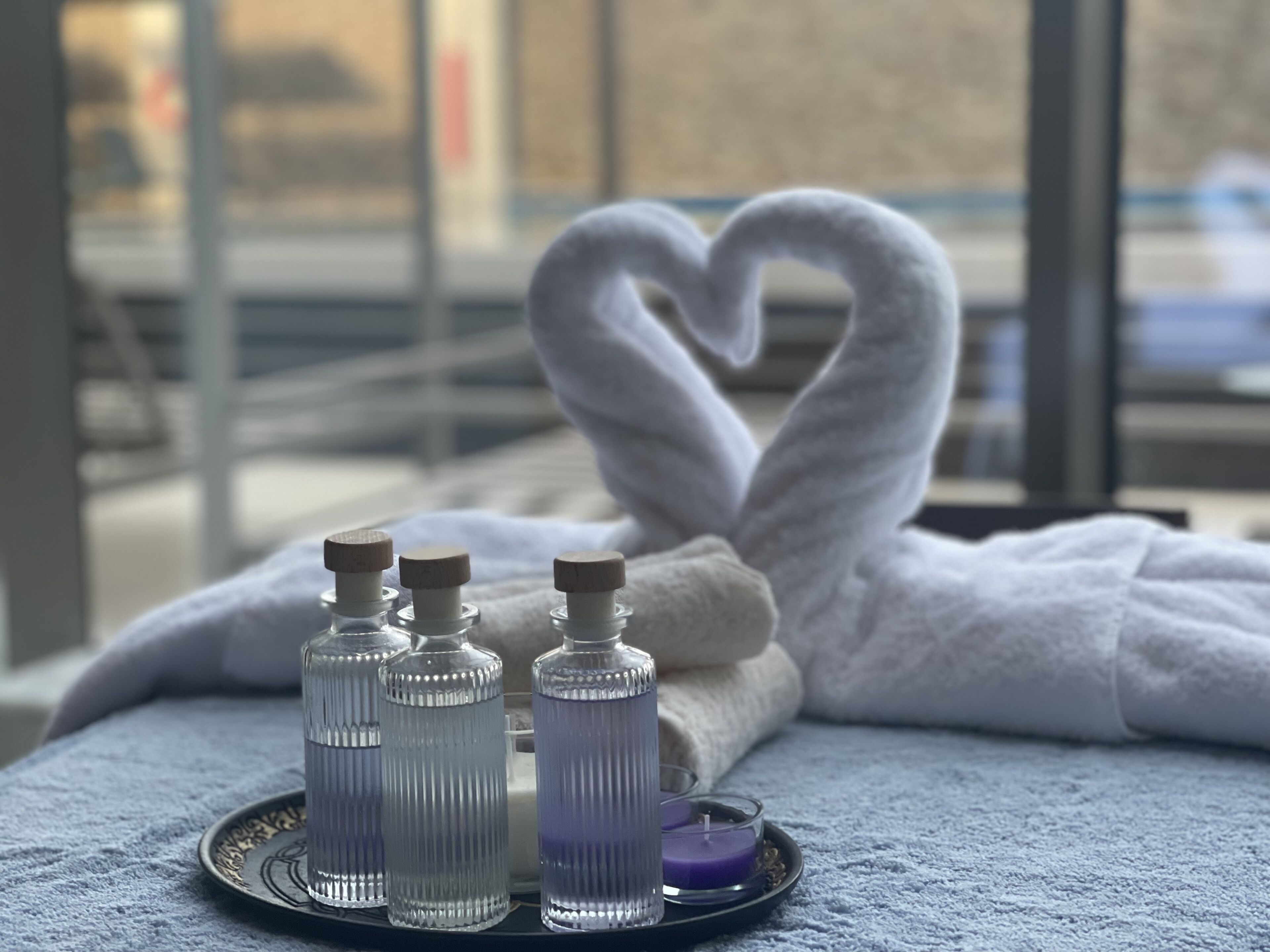 Un baño con una toalla con forma de corazón, tres botellas de artículos de aseo y una vela morada sobre una bandeja.