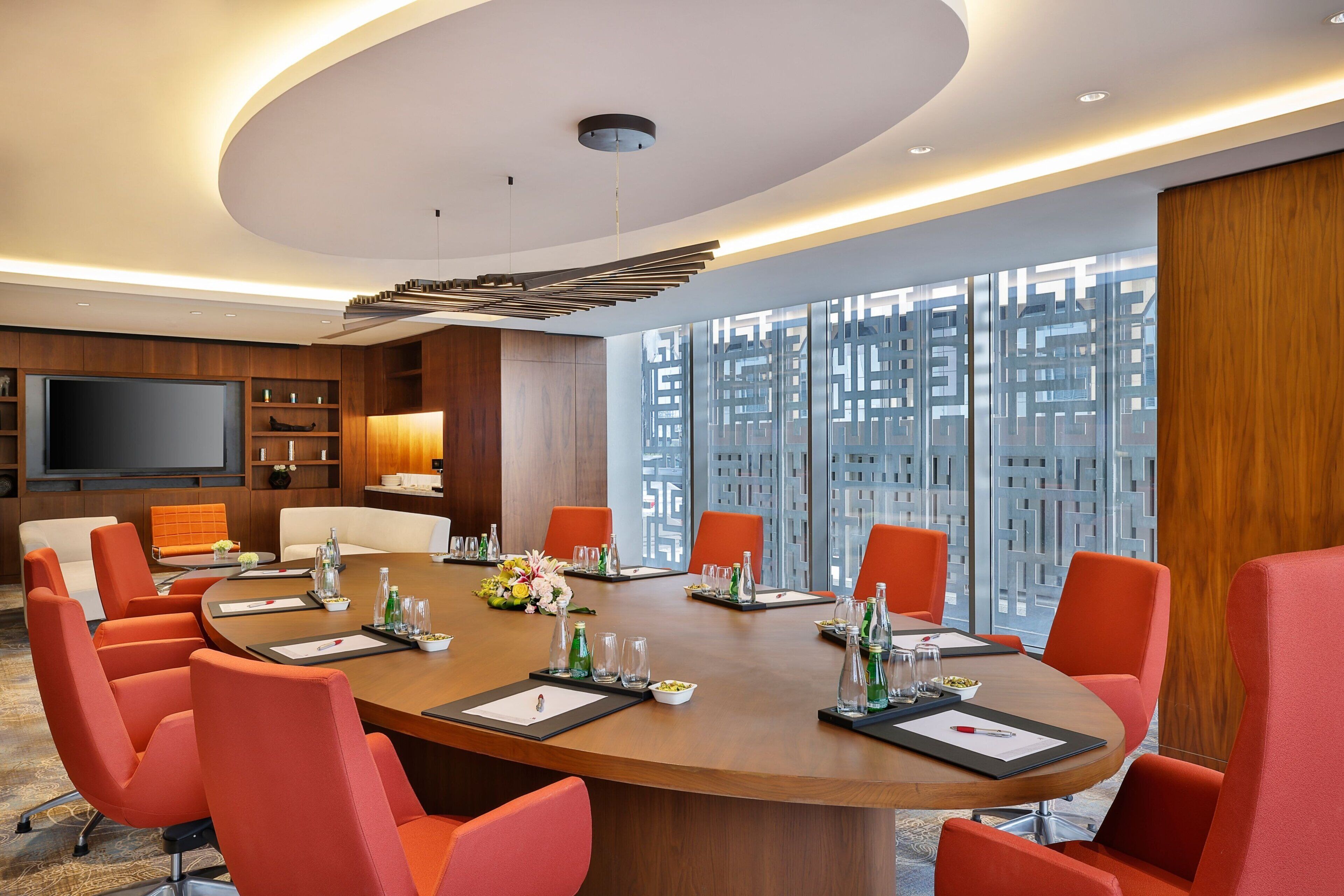 A modern conference room with a round table, orange chairs, and a large window with a geometric view.