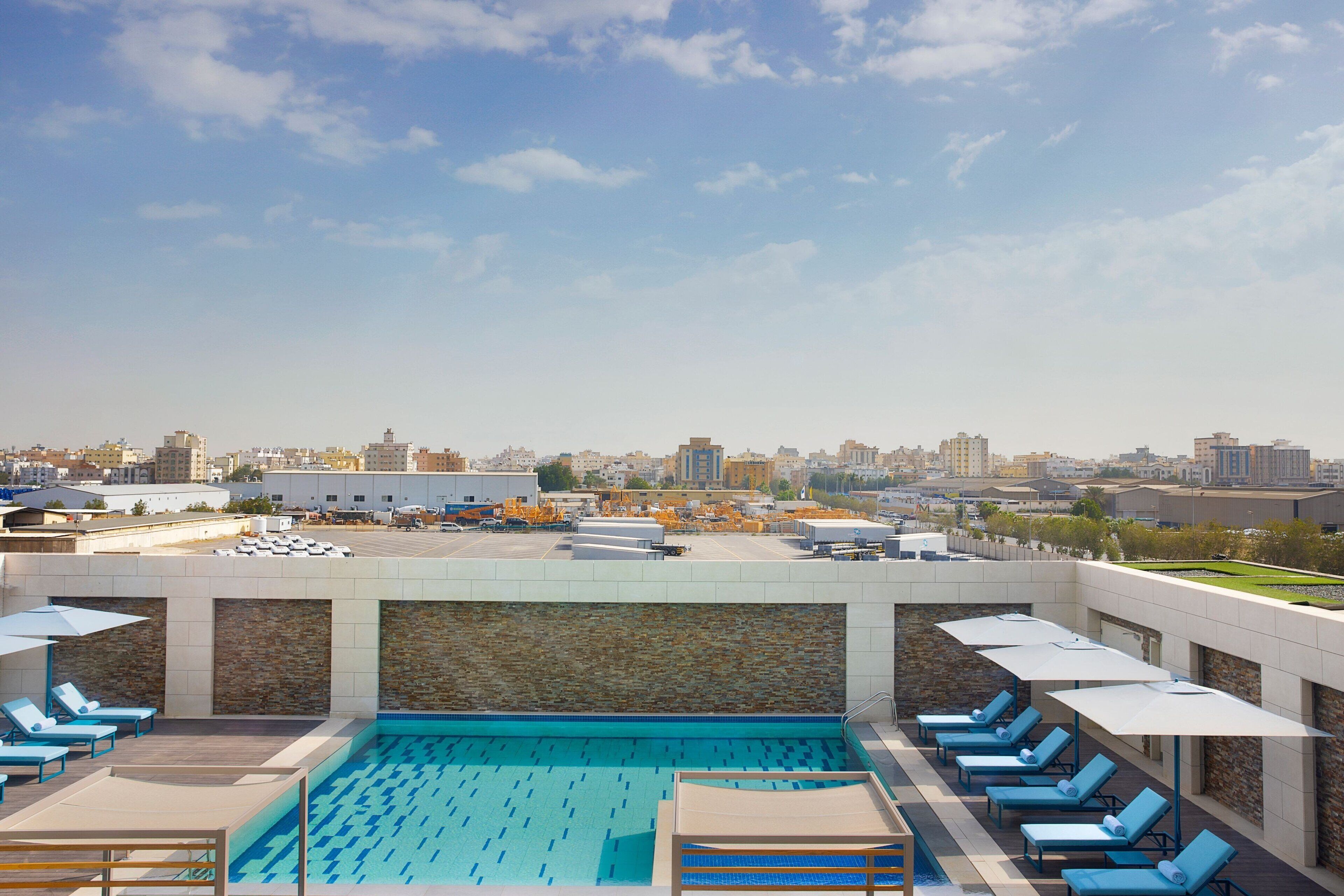 Une piscine sur le toit, agrémentée de chaises longues et de parasols, avec une vue sur la ville en arrière-plan et un ciel dégagé.