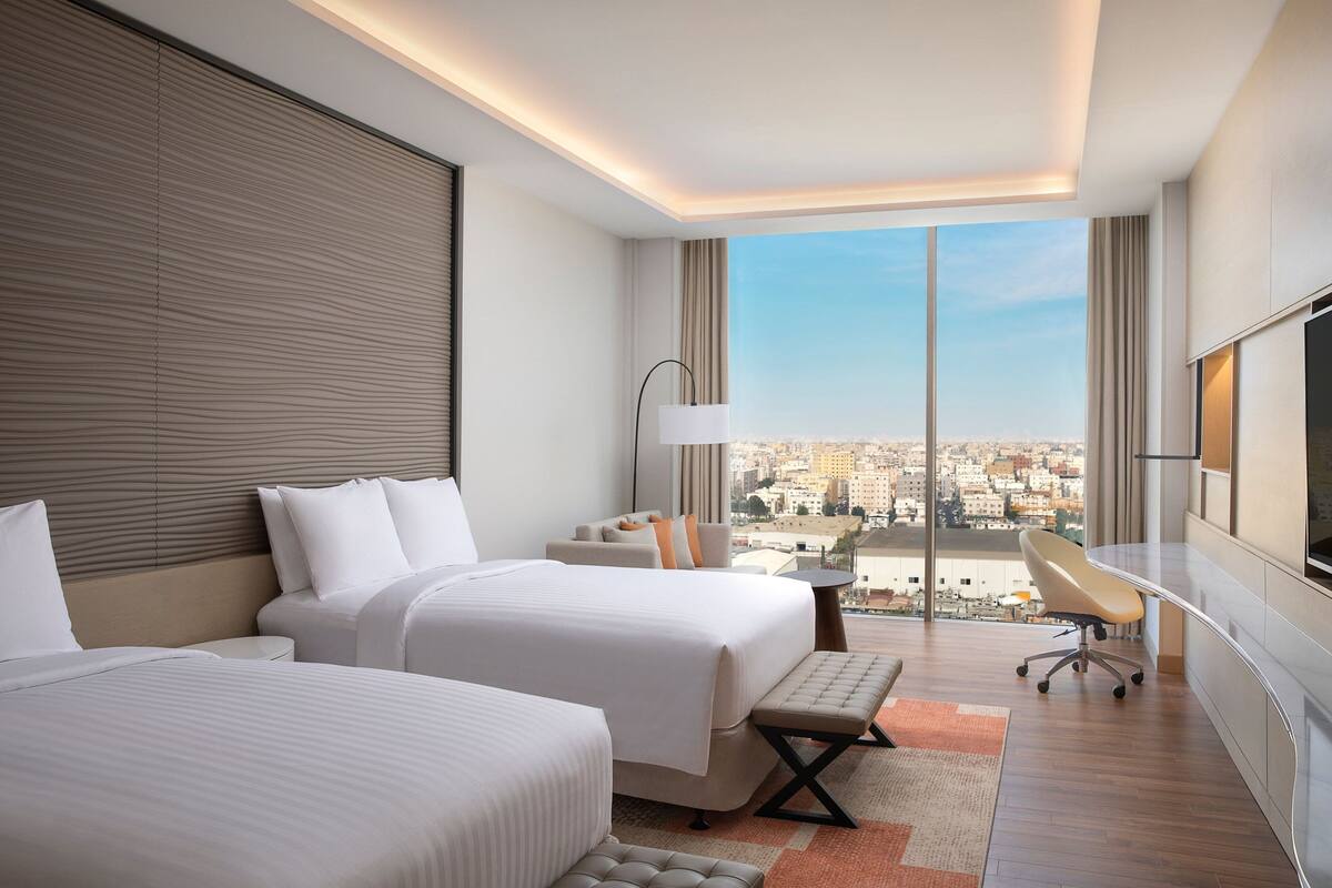 Une chambre d’hôtel moderne équipée de deux lits, d’un bureau, d’une chaise et d’une grande fenêtre donnant sur la ville.