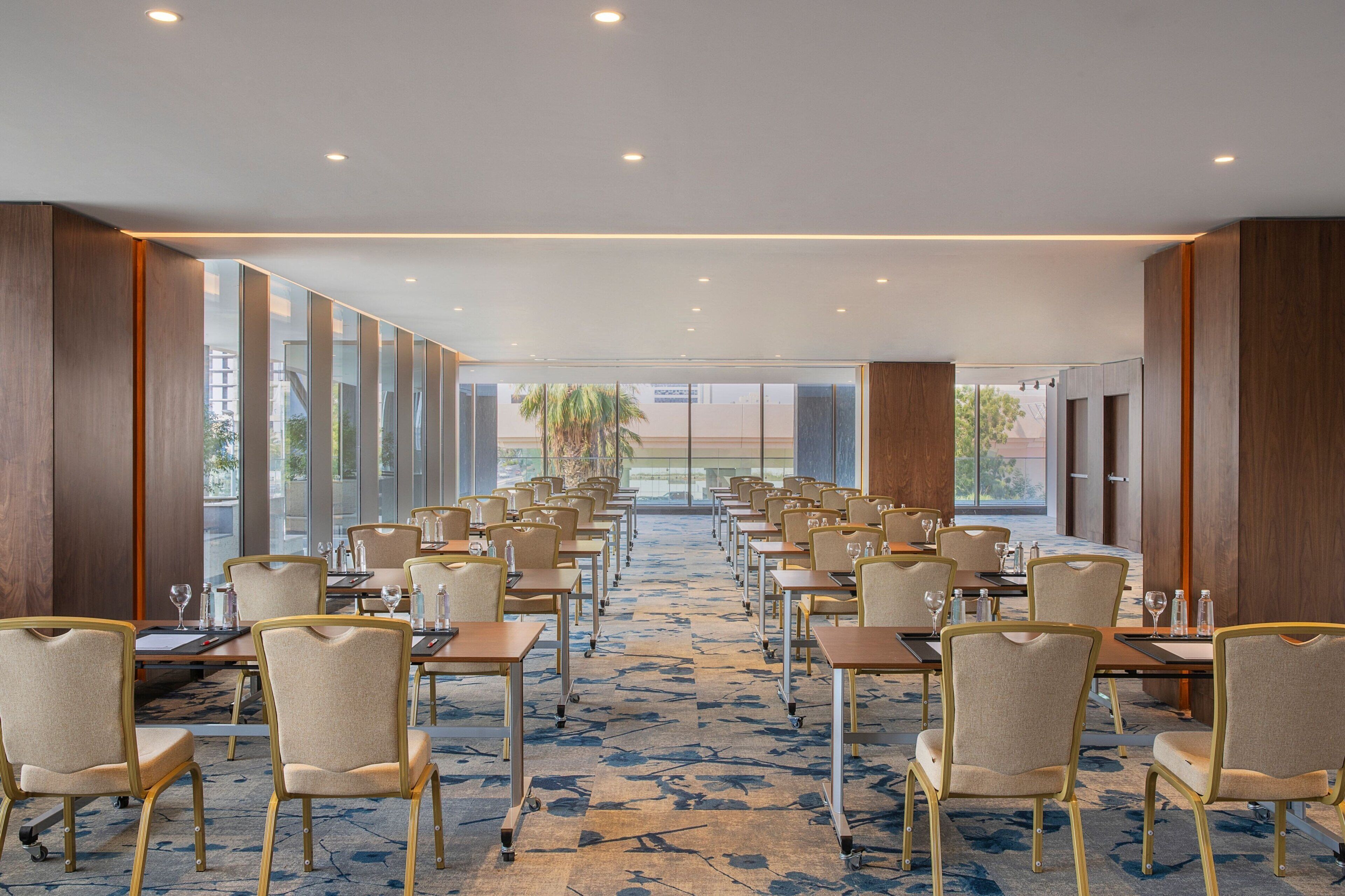 Un salón de conferencias con filas de mesas y sillas, ventanas amplias y piso alfombrado.
