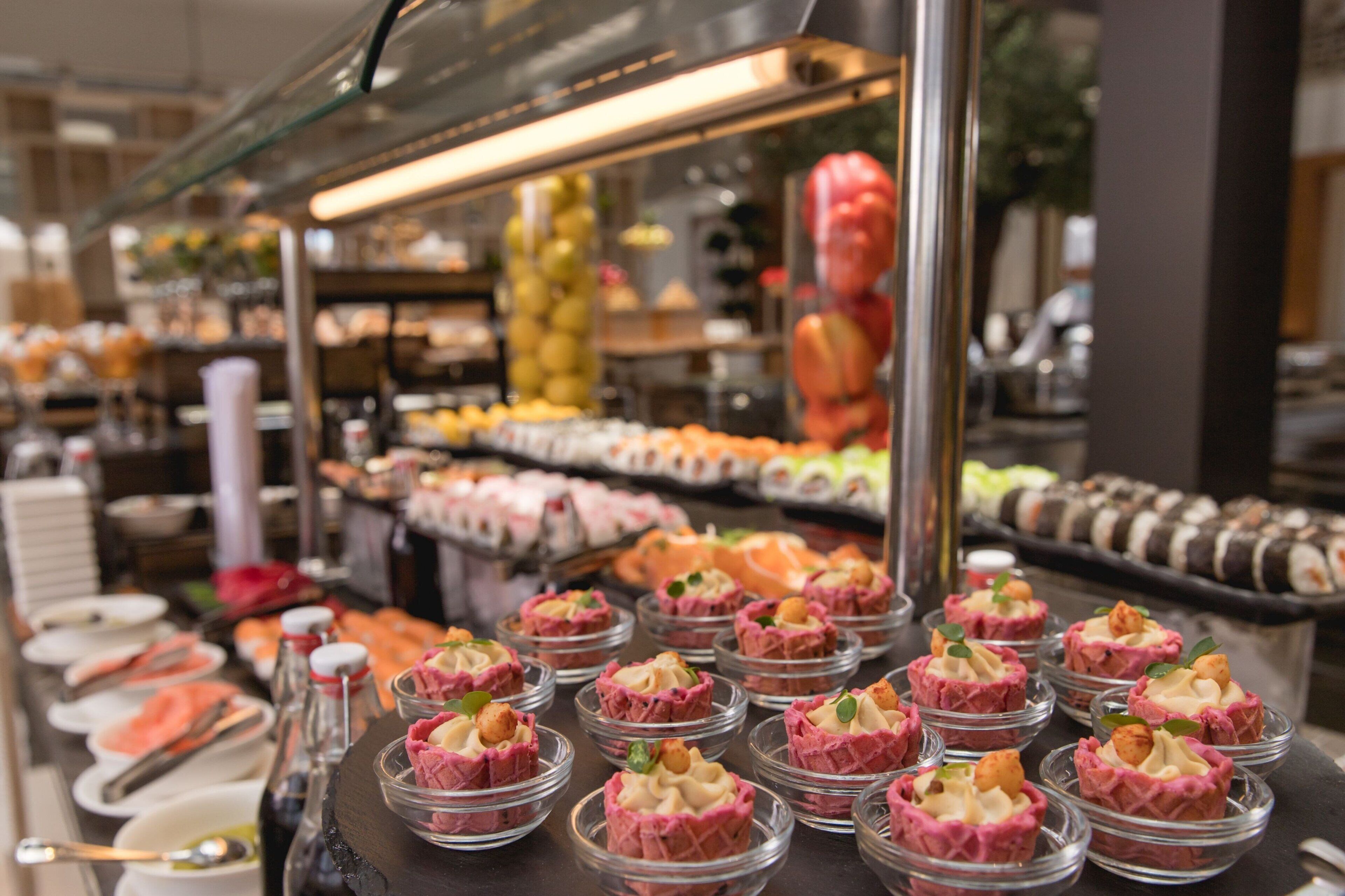 Un bufé con diversos alimentos, incluyendo una sección de tartaletas rosadas y una presentación de sushi.