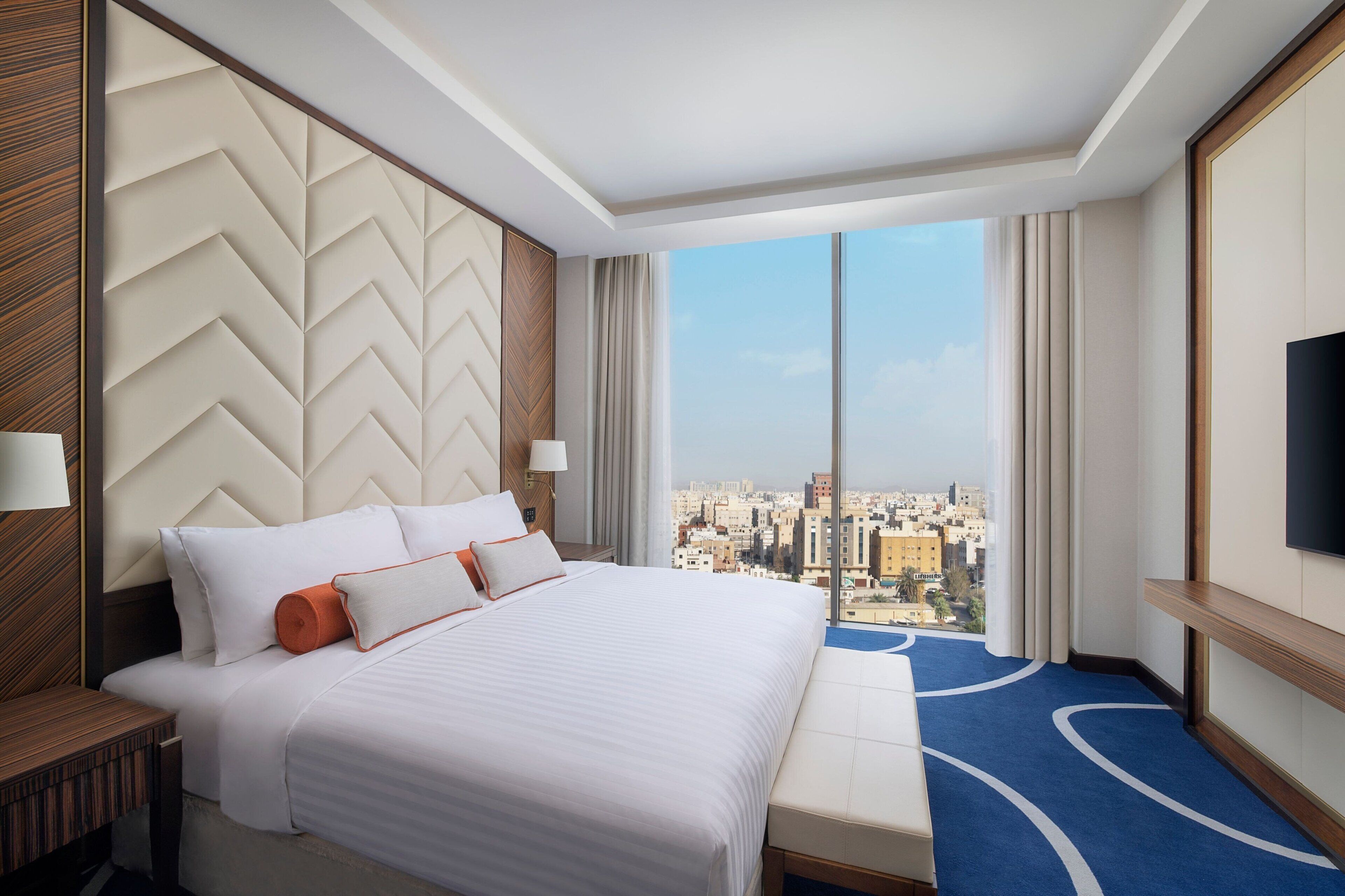 A hotel room with a large bed, a view of the city, and a flat-screen TV on the wall.