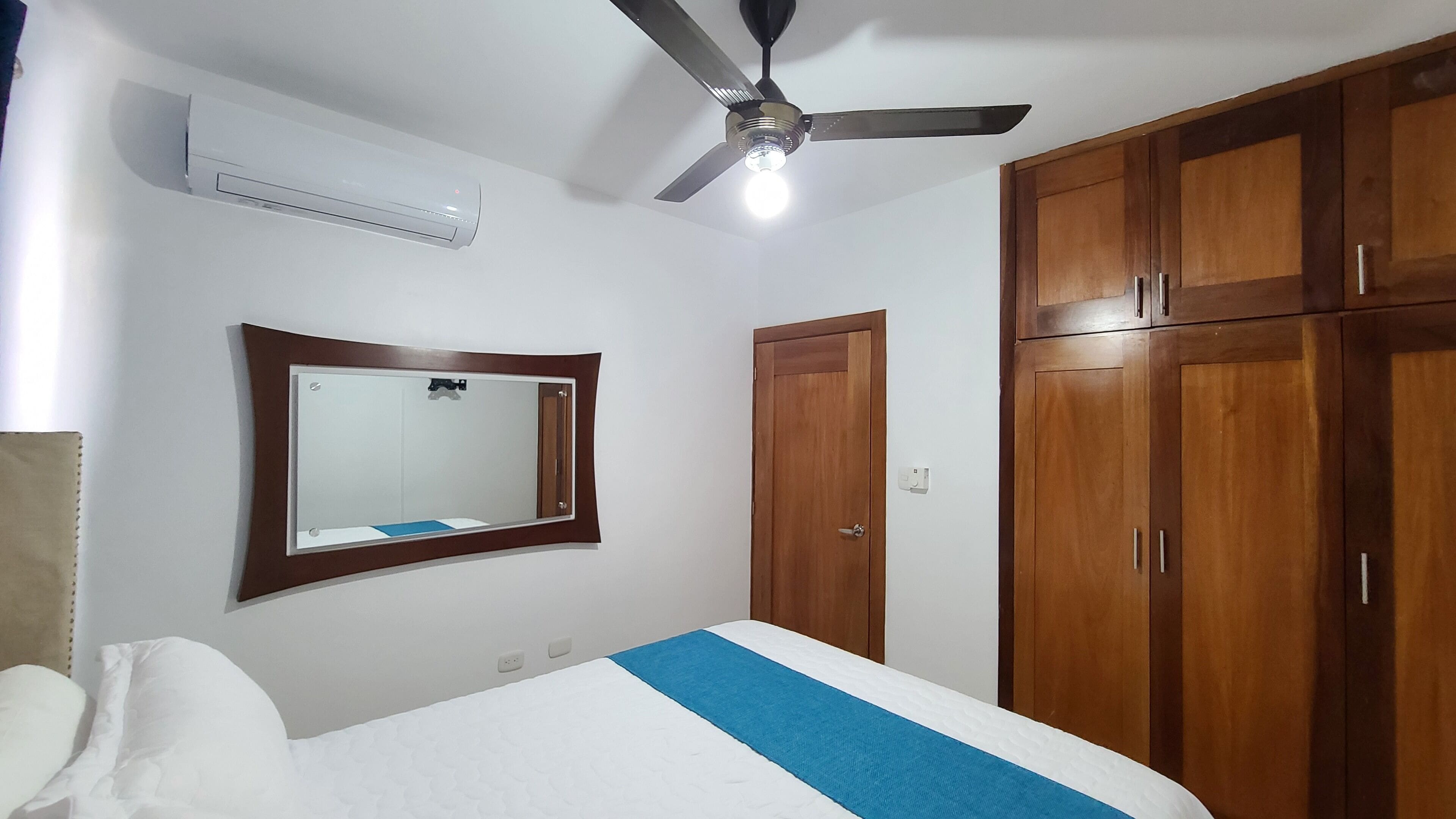 Un dormitorio con cama, ventilador de techo, aire acondicionado, espejo y armarios de madera.