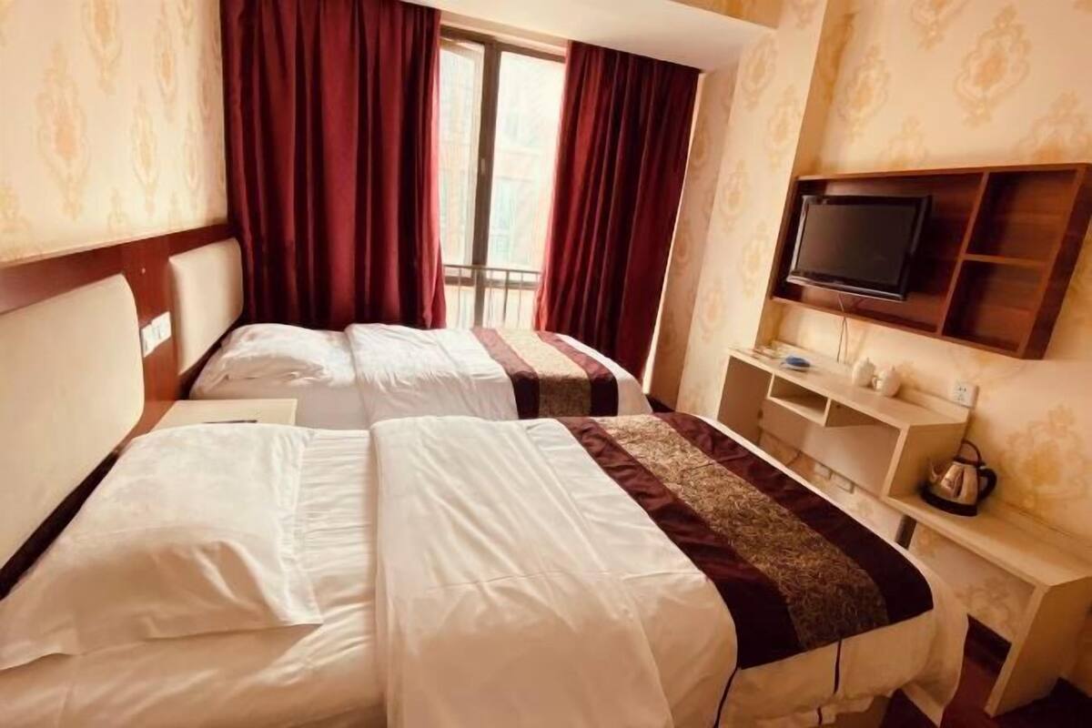 Ein Hotelzimmer mit zwei Betten, einem an der Wand montierten Fernseher und einem Fenster mit Vorhängen.