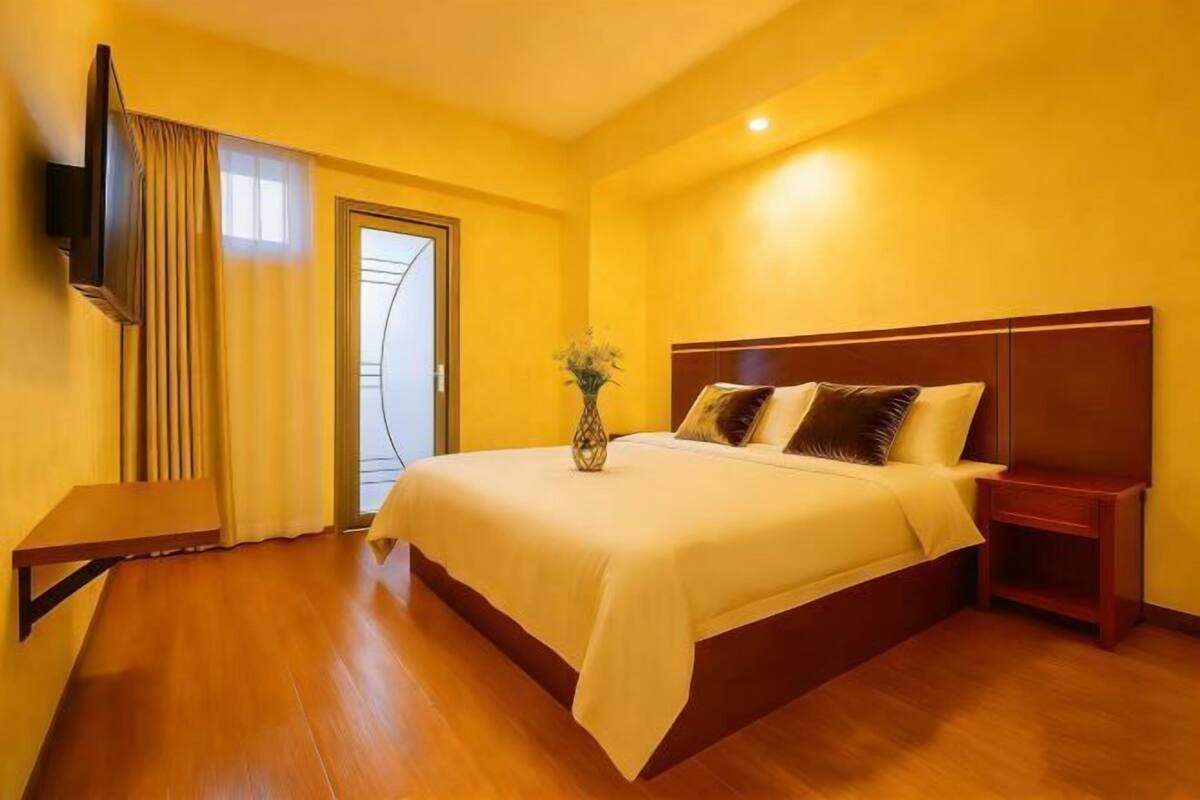 Ein Hotelzimmer mit einem großen Bett, einem Nachttisch, einem an der Wand montierten Fernseher und einem Fenster mit Vorhängen.