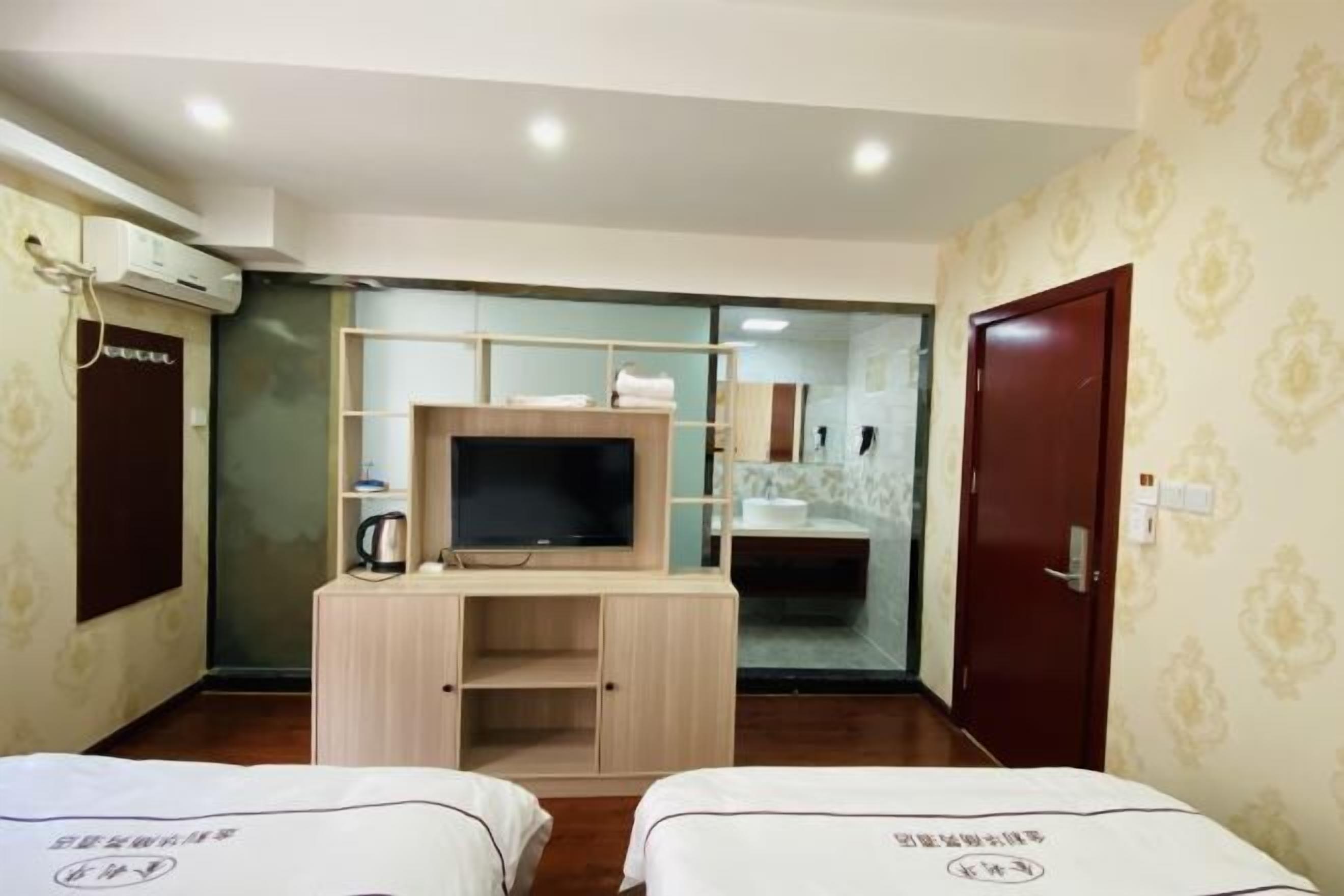 A hotel room with two beds, a TV, a desk, and a bathroom visible through a glass partition.