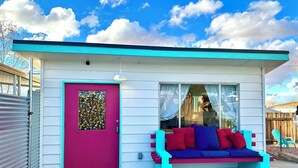Ein kleines Haus mit einer leuchtend pinken Tür, einer Sitzgelegenheit im Freien in Blau und Rot und einem großen Fenster.