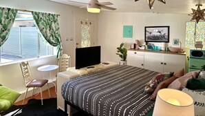 Ein Schlafzimmer mit Bett, Fernseher, Deckenventilator, Fenster mit Vorhängen, Schreibtisch, Stuhl und Kommode.