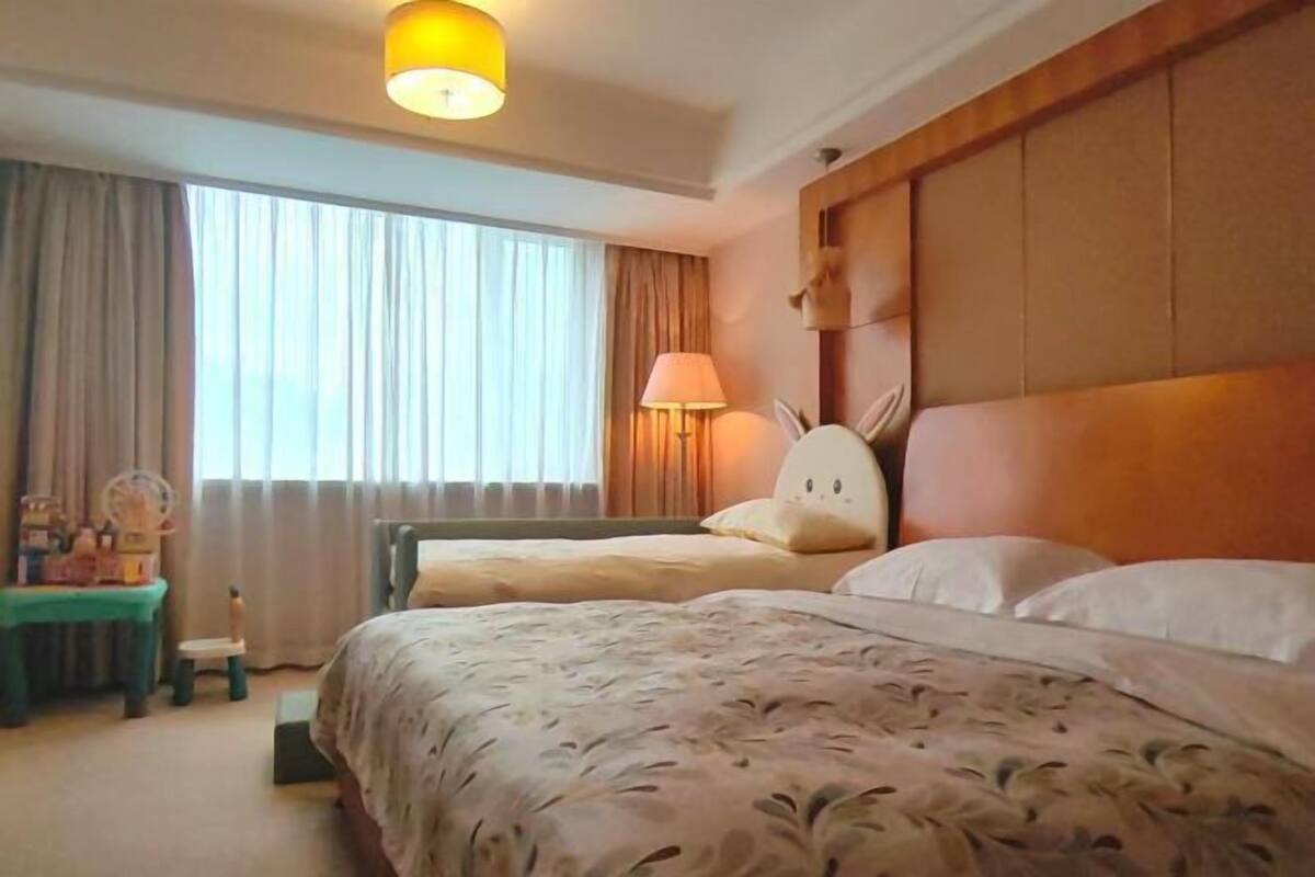 Habitació d'hotel amb un llit gran, un altre llit més petit, un capçal de llit de fusta, una taula de nit amb una làmpada i una finestra amb cortines.