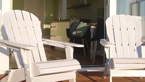 En terrasse med to hvide Adirondack-stole, en skydedør, der fører til et køkkenområde med et spisebord og stole, samt udsigt over naturen.