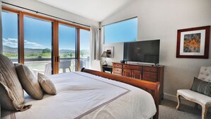 Ein Hotelzimmer mit einem großen Bett, einem Fernseher, einem Schreibtisch und einem Sessel. Große Fenster bieten Blick auf die Landschaft.