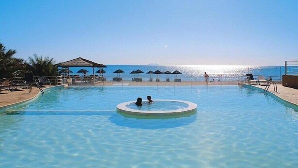 A clear swimming pool with a circular hot tub in the center, surrounded by lounge chairs and umbrellas, with a view of the sea in the background. - Apartment with terrace or balcony (Poggio-Mezzana)
