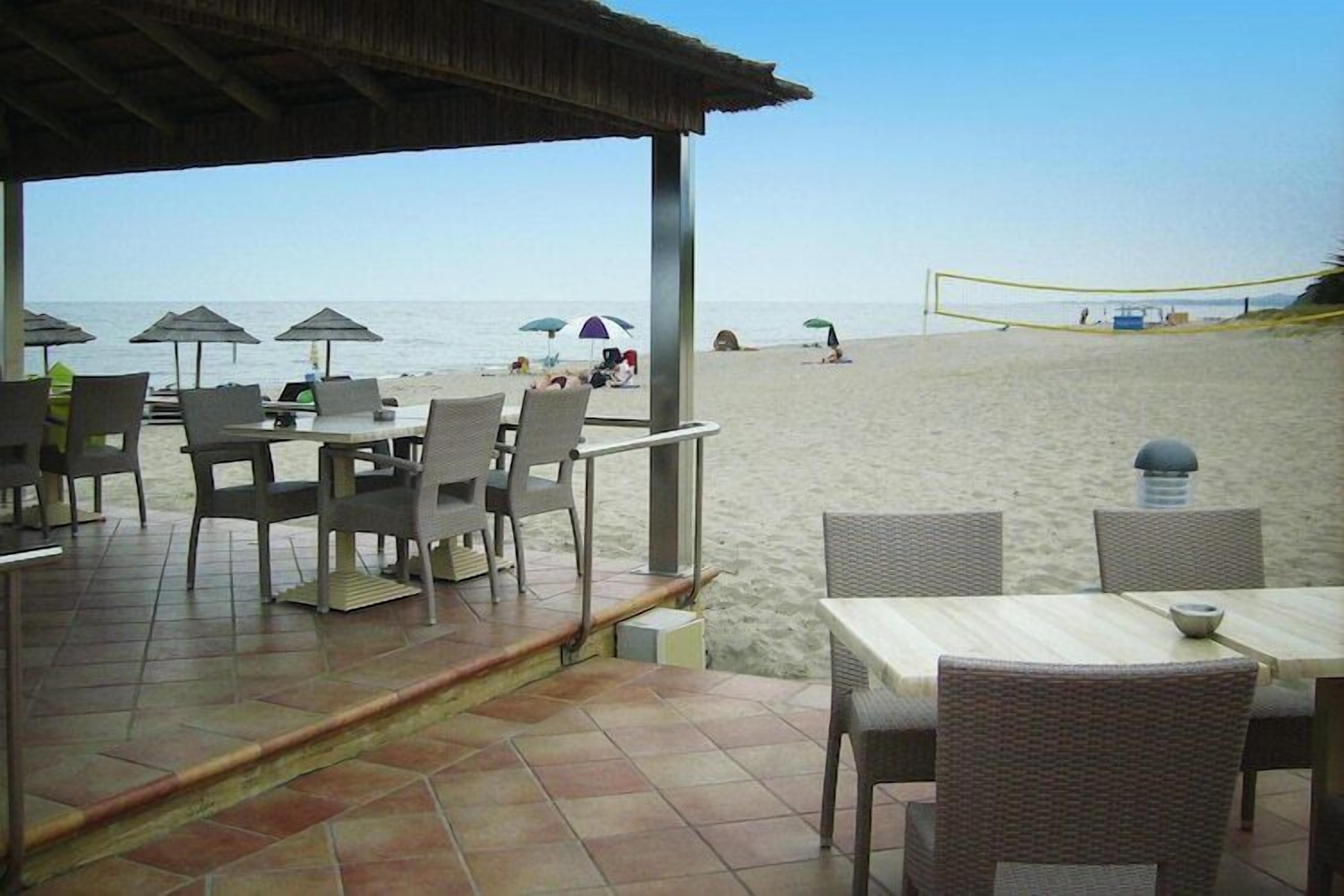 Un restaurante frente a la playa con mesas al aire libre, una red de voleibol y sombrillas.