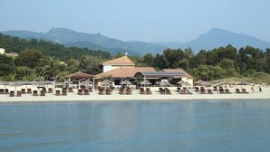 A beachside resort with sun loungers and umbrellas, a building with a red roof, and mountains in the background. - Apartment with terrace or balcony (Poggio-Mezzana)