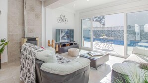 Ein modernes Wohnzimmer mit Sofa, Couchtisch und Fernseher. Ein großes Fenster bietet Blick auf einen Pool und eine Steinmauer.