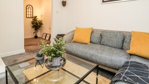 Een woonkamer met een grijze bank, gele kussens, een glazen salontafel en een potplant.