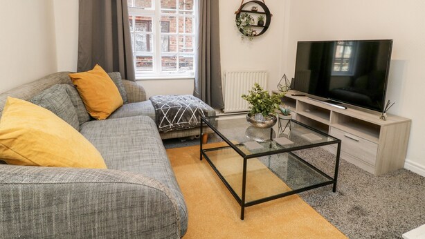 Een woonkamer met een grijze bank, gele kussens, een glazen salontafel, een tv op een dressoir en een raam met gordijnen.