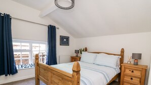 Een slaapkamer met een houten bed, een nachtkastje met lades, een raam met gordijnen, en een lijst met een afbeelding aan de muur.