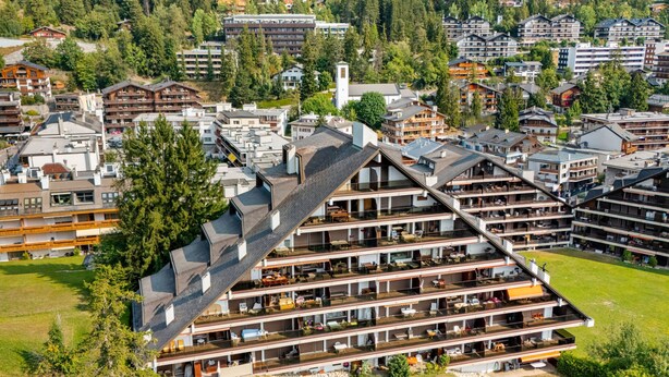 Un hôtel à plusieurs étages doté d’un toit incliné au design original, situé dans une région montagneuse, entouré de verdure et d’autres bâtiments.