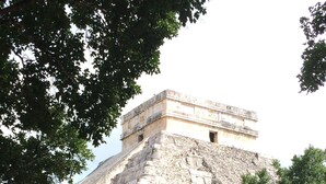 Una gran pirámide de piedra con cima plana, rodeada de un área verde con árboles y un banco.
