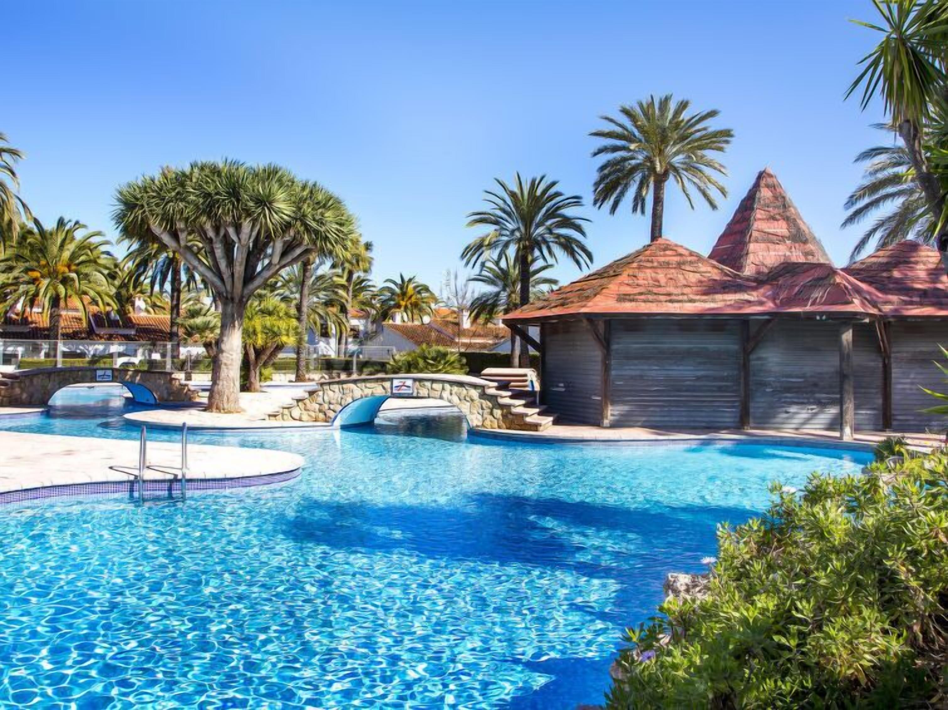 A swimming pool with a stone bridge, palm trees, and a building with a red roof.