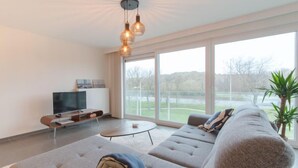 A modern living room with a large sectional sofa, a flat-screen TV, and a large window offering a view of the outdoors. - Ocean with parking by Interhome (Bredene)
