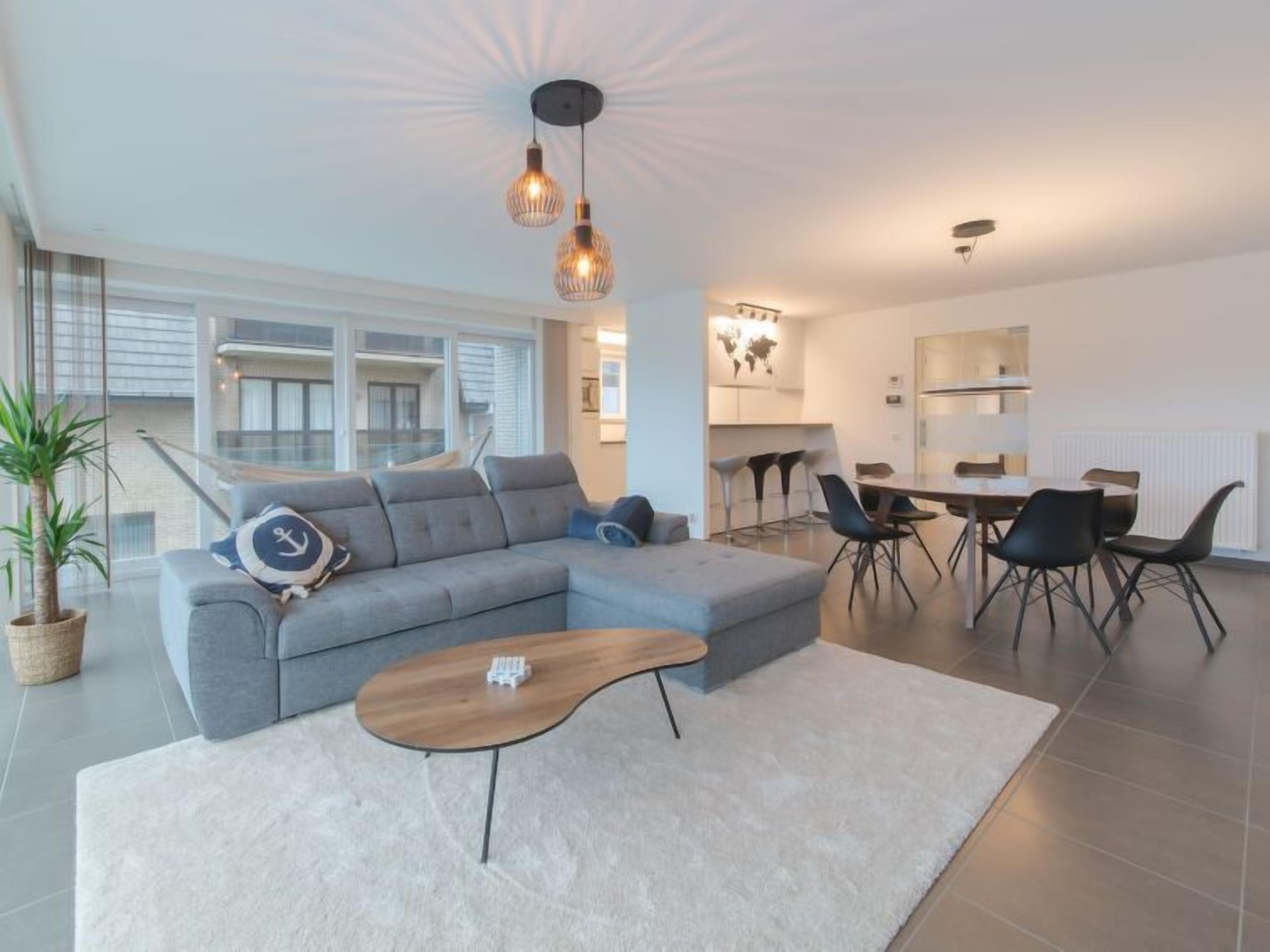 A modern living room with a grey sectional sofa, a wooden coffee table, and a dining area with a round table and black chairs.
