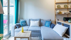 A modern living room with a grey sectional sofa, a wooden coffee table, and a white furry rug. There is a window with teal curtains, a bookshelf with decorative items, and a view of the outside balcony. - Les Normandières by Interhome (Cabourg)
