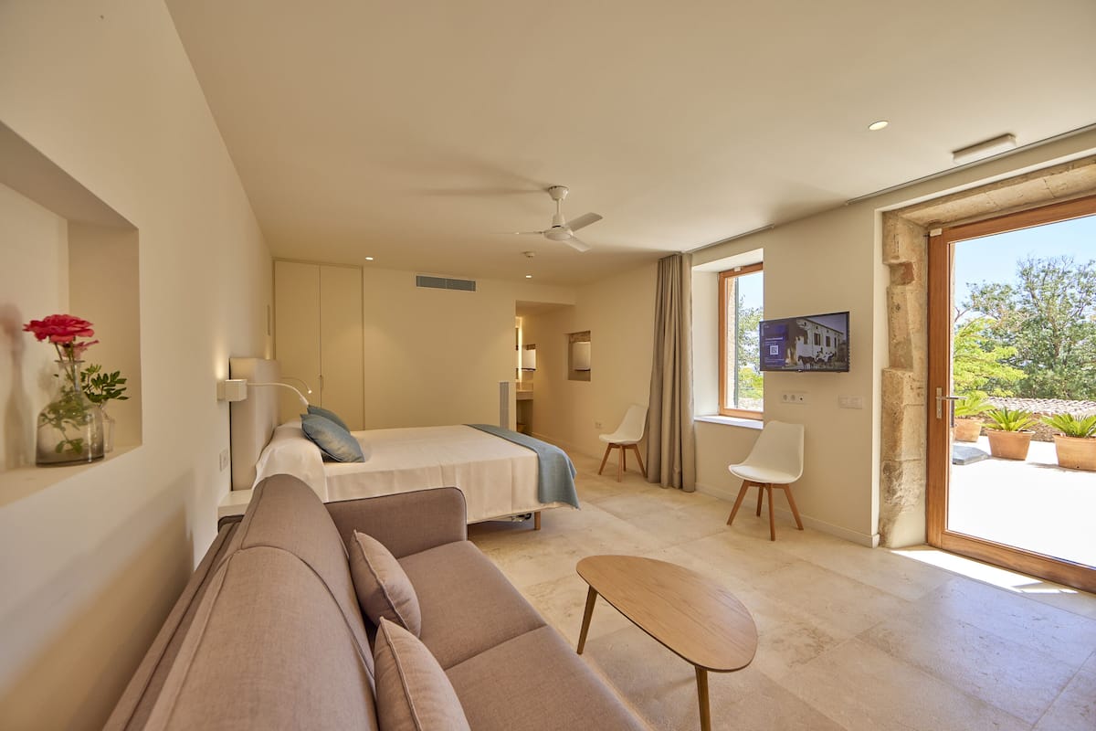 A modern hotel room with a bed, sofa, and a small table. There is a TV on the wall and a window with a view of greenery outside.