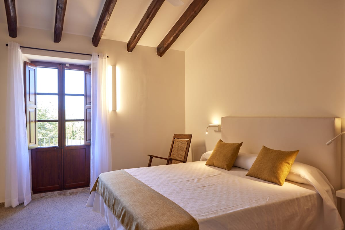 A bedroom with a large bed, a wooden chair, a door with curtains, and a wall-mounted light.