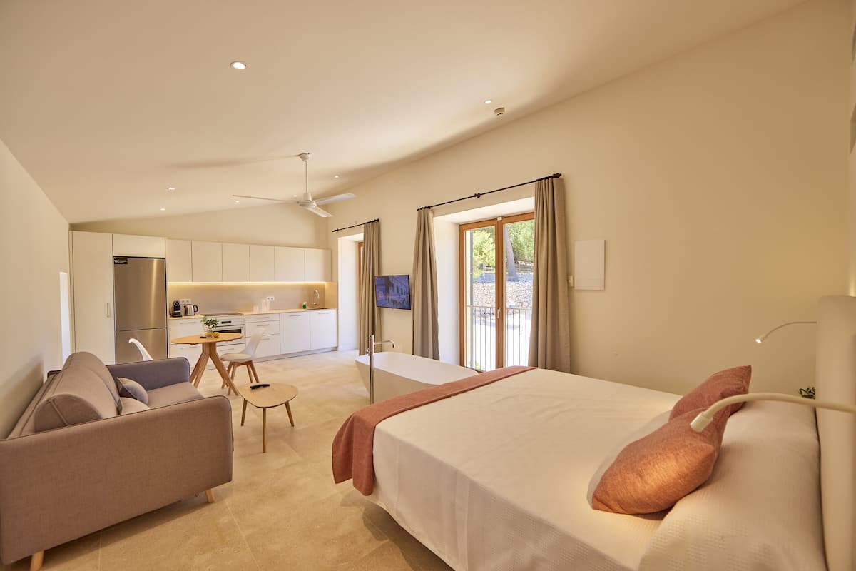 A modern hotel room with a large bed, a sofa, a dining area, and a kitchen.