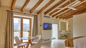 Une chambre d’hôtel avec un plafond en bois, un coin repas avec une table et des chaises, un lit recouvert d’une couverture marron, un téléviseur fixé au mur et une salle de bain avec un lavabo et un miroir.