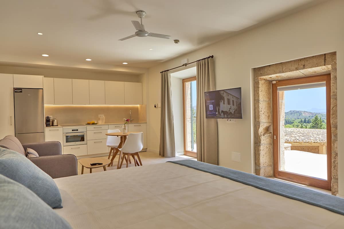 A modern hotel room with a bed, a small dining area, a kitchenette, and a large window offering a scenic view.